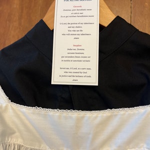 May include: A white tag with black text hanging on a black cassock and white surplice. The text reads "VESTING PRAYERS FOR ALTAR SERVERS" and includes prayers for the cassock and surplice.