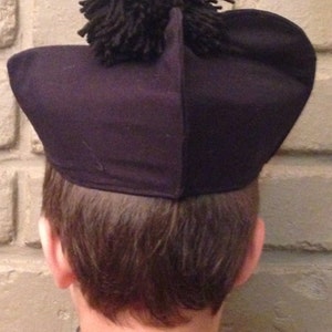 May include: A black felt hat with a large black pom-pom on top. The hat is worn by a person with brown hair.