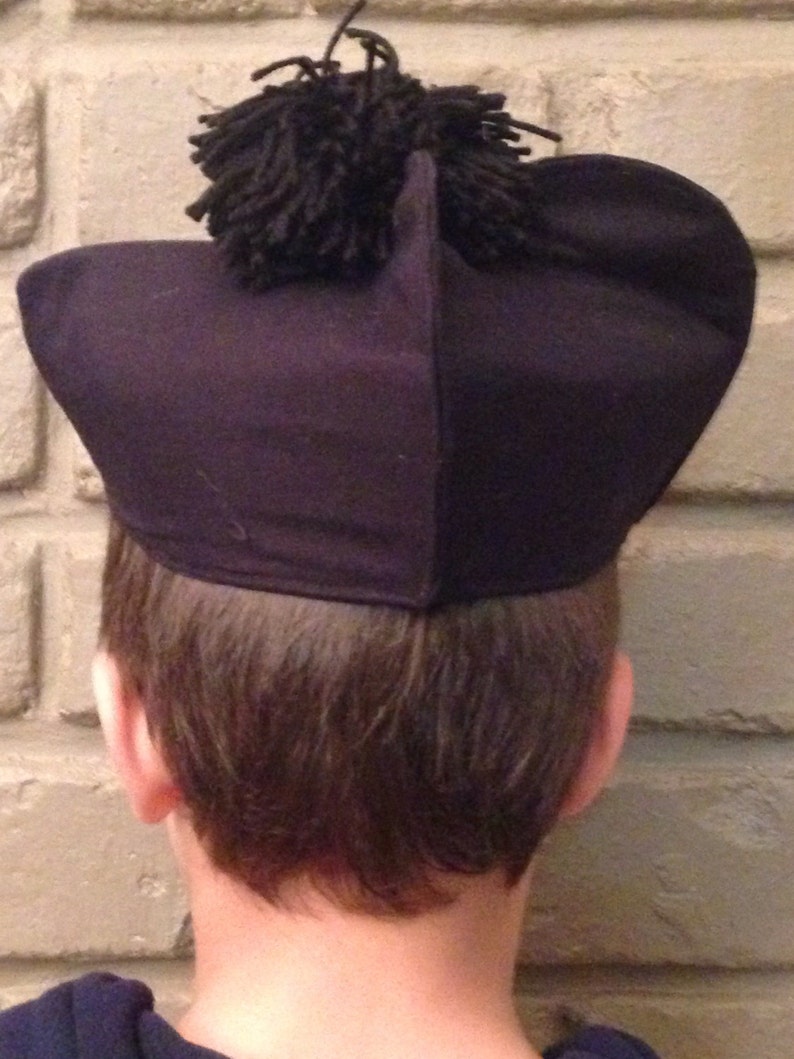 May include: A black felt hat with a large black pom-pom on top. The hat is worn by a person with brown hair.