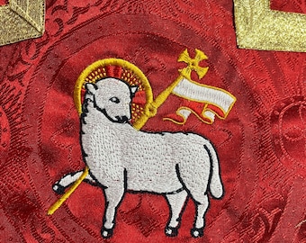May include: A red fabric with a gold border and an embroidered white lamb with a halo and a cross. The lamb is standing on a red background with a gold border.