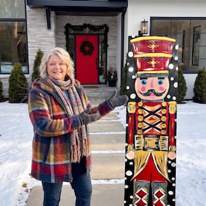 May include: A tall, rectangular wooden sign featuring a hand-painted nutcracker soldier in red, gold, black, and white. The soldier wears a tall hat, a uniform with gold accents, and stands against a black background decorated with white snowflakes.