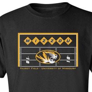 May include: Black t-shirt featuring a graphic of a football field with the word "MIZZOU" in yellow diamonds. Below the field is a tiger logo and the text "FAUROT FIELD - UNIVERSITY OF MISSOURI".