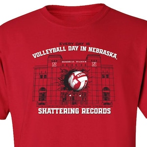 May include: Red t-shirt with a black and white graphic of a volleyball breaking through the wall of Memorial Stadium. The text "Every day is Volleyball Day in Nebraska. Shattering Records" is printed on the shirt.