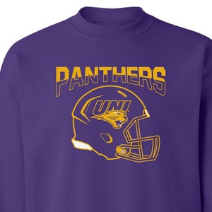 May include: A purple sweatshirt with the word "PANTHERS" in gold lettering above a gold football helmet graphic. The helmet has the letters "UNI" and a panther logo. The sweatshirt has a crew neck and long sleeves.