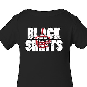 May include: Black baby bodysuit with the words "BLACK SHIRTS" in white block letters. A red and white football helmet graphic is in the center of the text. The bodysuit is short-sleeved and has snaps at the bottom.