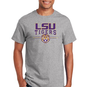 Puede incluir: Una camiseta gris con el logotipo de los LSU Tigers en morado y dorado. El logotipo presenta una cabeza de tigre con fondo morado y detalles dorados.