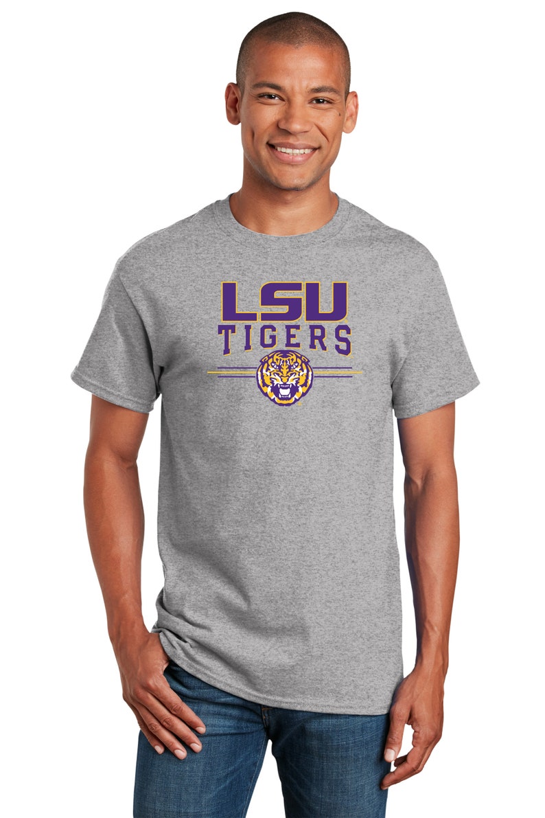 Puede incluir: Una camiseta gris con el logotipo de los LSU Tigers en morado y dorado. El logotipo presenta una cabeza de tigre con fondo morado y detalles dorados.