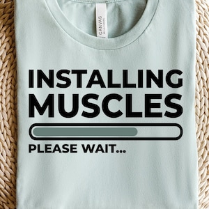 May include: A pale blue t-shirt featuring the text "INSTALLING MUSCLES" in black, with a loading bar graphic. Below the text is "PLEASE WAIT..." in black. The shirt is presented on a woven, circular surface.