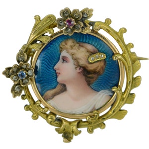 Könnte beinhalten: Eine goldfarbene Brosche mit einem blauen Emaille-Porträt einer Frau mit einem Rubin- und Diamant-Akzent. Die Brosche ist mit einem floralen Design verziert.