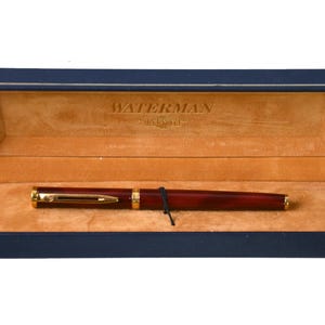 Pode incluir: Uma caneta-tinteiro Waterman vermelha e dourada em sua caixa de veludo azul. A caixa está aberta e a caneta está dentro. A caneta tem uma borda dourada e um design marmoreado vermelho e dourado.