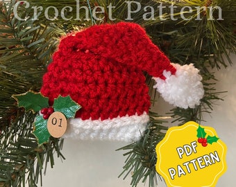 May include: Crocheted Santa hat ornament in red and white with a white pom-pom and a green holly sprig with red berries. The ornament has a brown tag with the number "01" on it. The ornament is hanging on a green pine tree branch. The text "PDF PATTERN" is on a yellow circle with a green holly sprig with red berries.