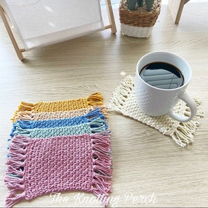 May include: Five crocheted coasters in pink, green, blue, beige, and yellow with fringe edges. The coasters are arranged in a stack on a wooden table. A white coffee mug with a black coffee sits on a crocheted coaster.
