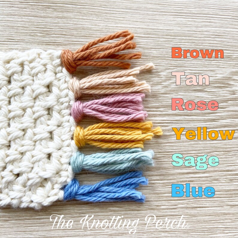 Pu&ograve; includere: Primo piano di un pezzo di uncinetto bianco con frange colorate. Le frange sono di colore marrone, beige, rosa, giallo, salvia e blu. Il testo "The Knotting Perch" &egrave; visibile nella parte inferiore dell'immagine.