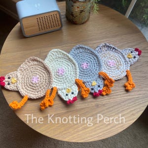 May include: Crocheted chicken-shaped coasters in various colors, including beige, white, and gray. Each coaster features a pink flower in the center and orange feet. The text "The Knotting Perch" is visible.