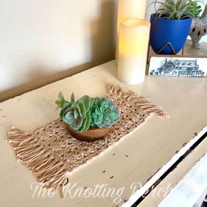 May include: A small, beige crocheted placemat with fringe, holding a wooden bowl filled with faux succulents. The text "The Knotting Porch" is visible at the bottom. The scene includes candles and decorative items.