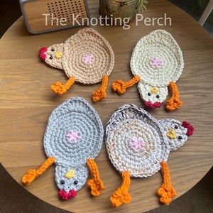 Puede incluir: Cuatro posavasos de ganchillo hechos a mano con forma de pollo, en beige, blanco, gris y una mezcla de gris y beige. Cada posavasos tiene un cuerpo circular, una cabeza de pollo y patas naranjas. El texto "The Knotting Perch" es visible.