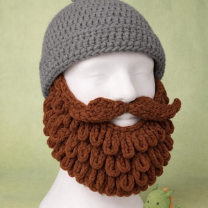 May include: A gray crocheted beanie hat with a matching brown crocheted beard and mustache set. The beard and mustache are styled with a curly design. The set is displayed on a white mannequin head.
