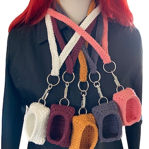 May include: Six crocheted phone cases in various colors, including white, burgundy, orange, gray, and pink, are attached to crocheted lanyards. The lanyards are crossed and hanging from a mannequin's neck.