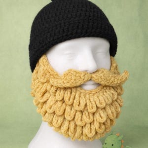 May include: A black crocheted beanie hat with a matching yellow crocheted beard and mustache set. The beard has a scalloped design. The hat has a small black pom-pom on top. The set is displayed on a white mannequin head.