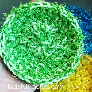 May include: Green and white crocheted scrubby with a fluffy texture. The scrubby is round and has a center hole. KidsNKcrochet.com is visible in the image.