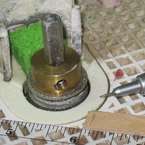 May include: Close-up of a metal and brass tool assembly with a silver drill bit. The assembly includes a hexagonal metal piece, a brass ring, and several silver-colored discs. A green sponge is visible.