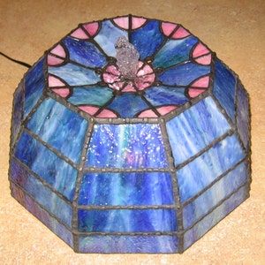 May include: A stained glass lampshade with a blue and pink geometric design. The lampshade has a central opening with a clear glass piece in the center.