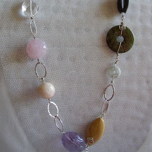 May include: A silver chain necklace with a variety of gemstone beads, including pink, brown, green, and purple. The necklace also features a silver circular pendant with a decorative design.