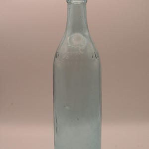 May include: A clear glass bottle with a rounded bottom and a narrow neck. The bottle is light blue in color and has a faint inscription on the side.