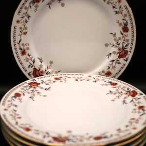 May include: White ceramic plates with a floral design in red, blue, and gold. The plates have a gold rim and a repeating pattern around the edge. The plates are stacked, with one plate on top.