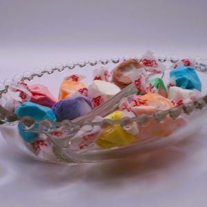 May include: A clear glass bowl with a beaded rim filled with colorful candies wrapped in white paper. The candies are in various shapes and sizes.