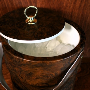 May include: A brown faux wood ice bucket with a white interior and a gold-colored lid with a ring handle. The bucket is filled with ice cubes.