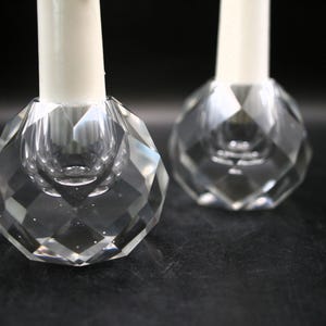 May include: Two clear, faceted crystal candle holders with white candles. The candle holders have a geometric design and sit on a dark surface. The focus is on the crystal's intricate cut and the contrast with the white candles.