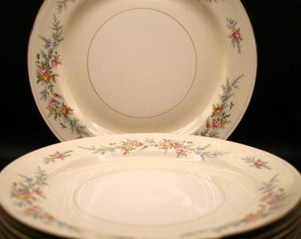 May include: A stack of cream-colored plates with a floral design. Each plate features a gold rim and a delicate pattern of pink, yellow, and blue flowers. The plates are stacked on a black surface.