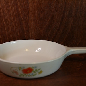 May include: White ceramic skillet with a single handle. The skillet has a floral design with red, orange, yellow, and blue flowers. The skillet is on a dark brown wooden surface.
