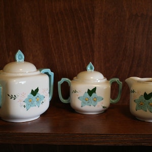May include: A white teapot, sugar bowl, and creamer set with blue and green floral designs. The teapot has a square body and a blue lid. The sugar bowl and creamer have round bodies.