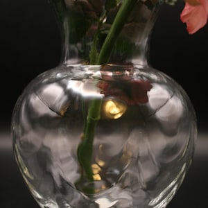 May include: A clear glass vase with a textured, rippled surface. A green stem and a pink rose are visible inside the vase. The vase has a rounded base and a flared top, set against a dark background.