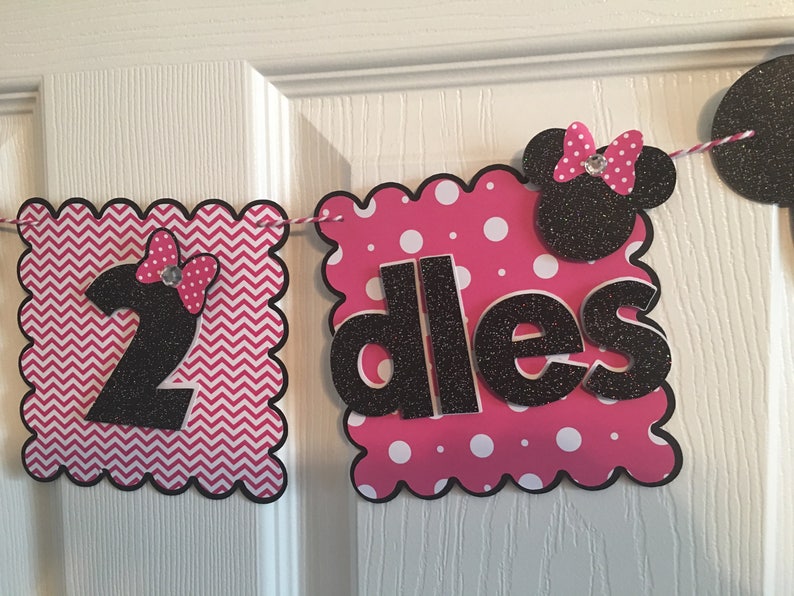 May include: A pink and black banner with a white chevron pattern and white polka dots. The banner says "2 dlles" with a black Minnie Mouse head in the middle.