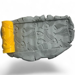 May include: A gray, rectangular clay slab with fish skeleton impressions. A yellow roller with fish skeleton designs is positioned on the left side. The clay slab has a rough, textured surface and a white background.