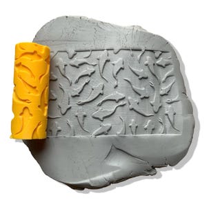May include: A yellow textured roller next to a gray clay slab. The roller has a repeating pattern of curved shapes. The clay slab is imprinted with a pattern of fish and other aquatic shapes, creating a textured surface.