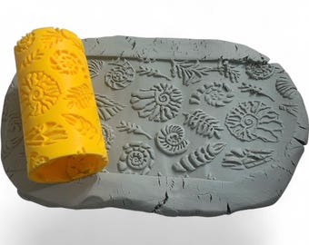 May include: A yellow rolling pin with a fossil pattern next to a gray clay slab with fossil imprints. The rolling pin has a repeating pattern of spiral fossils. The clay slab has various fossil designs.