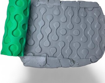 Può includere: Un rullo verde a motivi accanto a una lastra di argilla grigia. Il rullo presenta un motivo ripetuto di forme arrotondate collegate. La lastra di argilla ha lo stesso motivo impresso sulla sua superficie, creando un design strutturato.