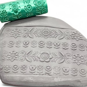 Puede incluir: Un rodillo verde con un patrón floral descansa sobre una superficie de arcilla gris, que también presenta un diseño floral en relieve. El rodillo es cilíndrico y tiene un patrón repetitivo de flores y hojas.