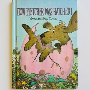 May include: A vintage children's book titled "How Fletcher Was Hatched!" features a whimsical illustration of a dog emerging from a pink egg. The cover has a light blue background with the author's names, Wende and Harry Devlin.