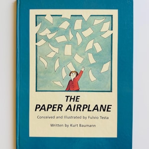 May include: A hardcover book titled The Paper Airplane, conceived and illustrated by Fulvio Testa and written by Kurt Baumann. The cover features a teal border and a central illustration of a person in a red shirt reaching toward falling papers.