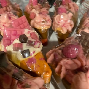 May include: Assortment of pink and brown chocolate-covered treats wrapped in clear plastic. The treats are shaped like cones and topped with chocolate squares and pink swirls.