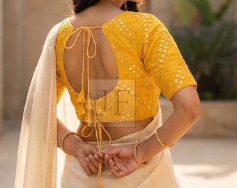 May include: A bright yellow blouse featuring intricate embroidery and small circular mirrors, paired with a cream-coloured fabric drape. The blouse has a deep open back design with thin yellow tie-up strings and decorative tassels hanging down.