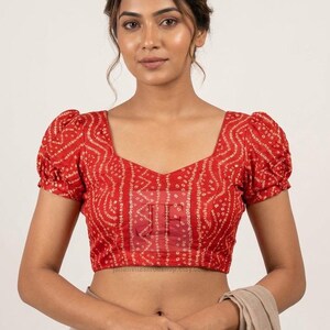 Könnte beinhalten: Eine rote Bluse mit weißem Bandhani-Tie-Dye-Muster, herzförmigem Ausschnitt und kurzen Puffärmeln. Das Kleidungsstück ist taillenkurz geschnitten und weist in der Mitte des Brustbereichs ein halbtransparentes Wasserzeichen-Logo auf.