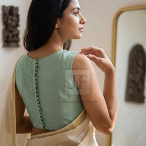 May include: A sleeveless sage green blouse featuring a vertical row of matching fabric-covered buttons running down the center back. The garment is paired with a cream-colored fabric draped over the shoulder. The text JF is visible in the center.