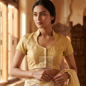 Op de afbeelding: Een goudkleurige blouse met korte mouwen, een opstaande kraag en een knoopsluiting aan de voorzijde, gecombineerd met een crèmekleurige sari met gouden ruitpatronen. De blouse heeft een gestructureerde pasvorm en een subtiele glans.