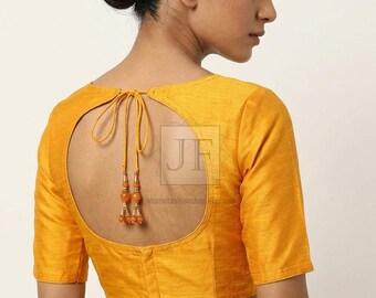 May include: A mustard yellow blouse with short sleeves and a large circular open back design. The back features a thin tie closure at the top with two dangling orange beaded tassels. The fabric has a subtle horizontal texture throughout.